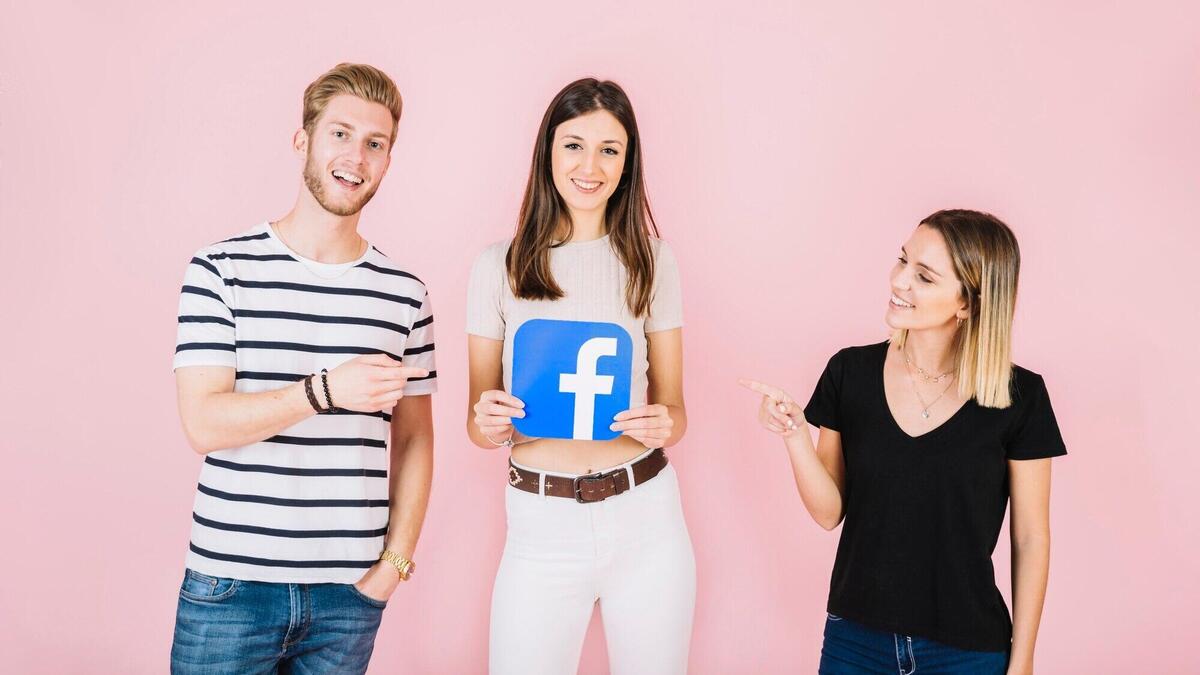 Man and woman pointing at their friend holding facebook icon on pink backdrop
