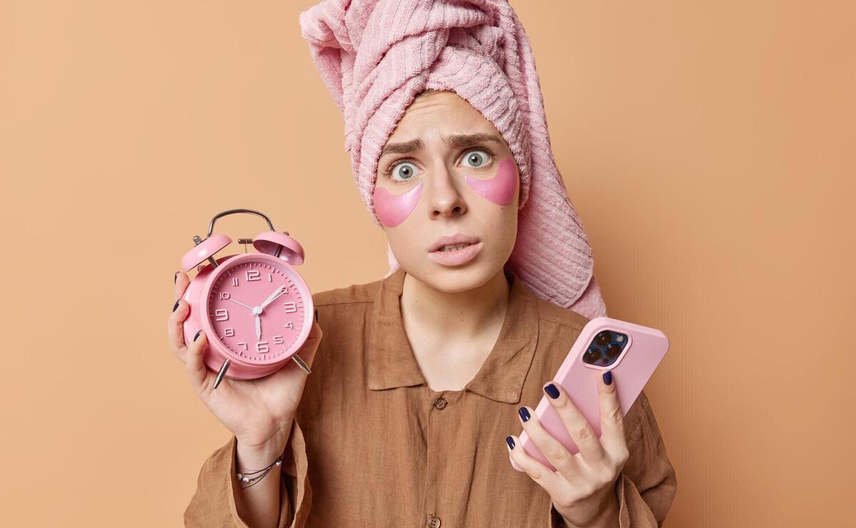 Worried young woman awakes late holds alarm clock and smartphone undergoes morning routines dressed in pajama wrapped towel on head isolated over brown background awakening and rest concept