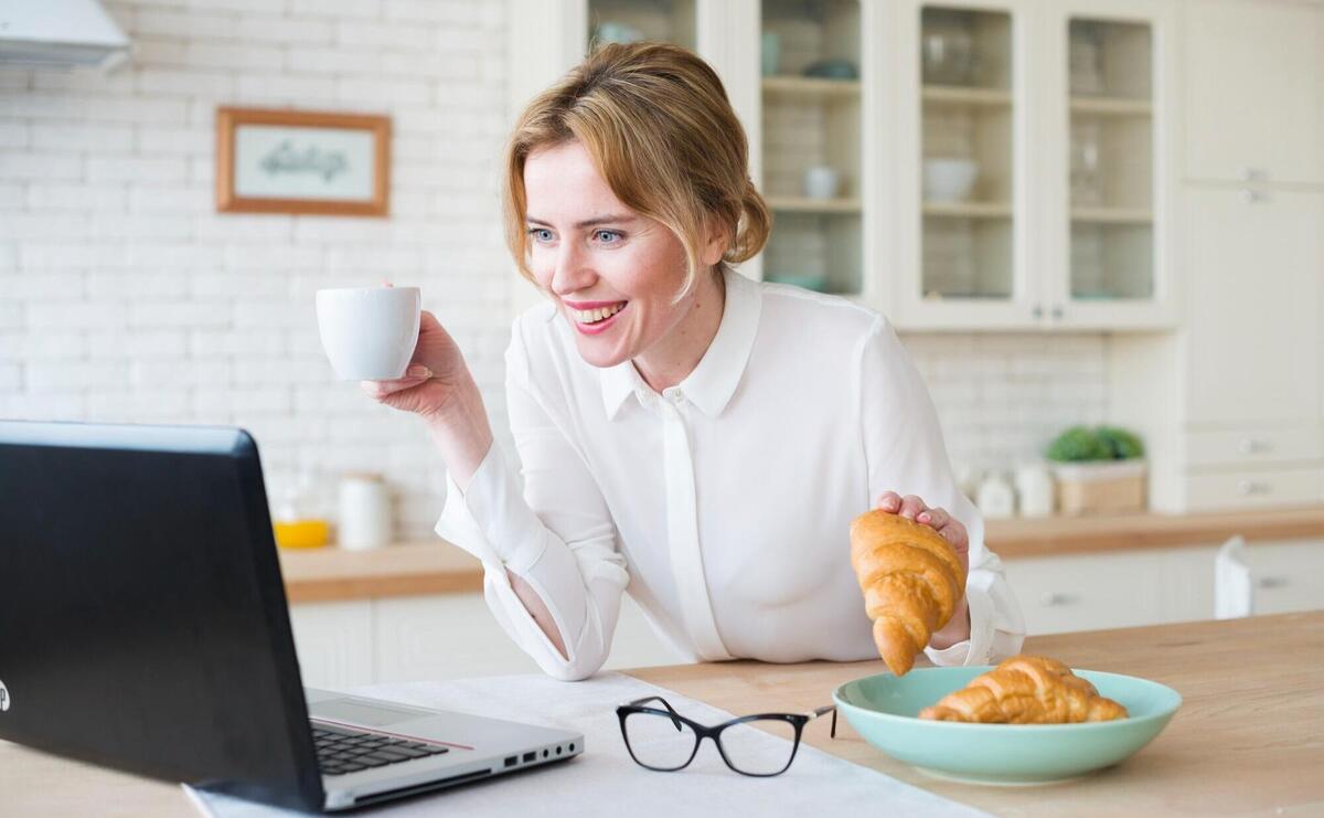Woman in white shirt with croissant using laptop.
