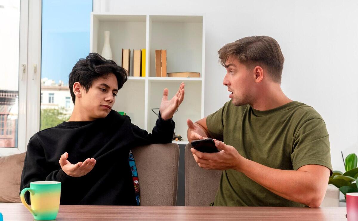 Unpleased young blonde handsome man holds and points at phone with hand sitting at table and looking at clueless young brunette handsome boy